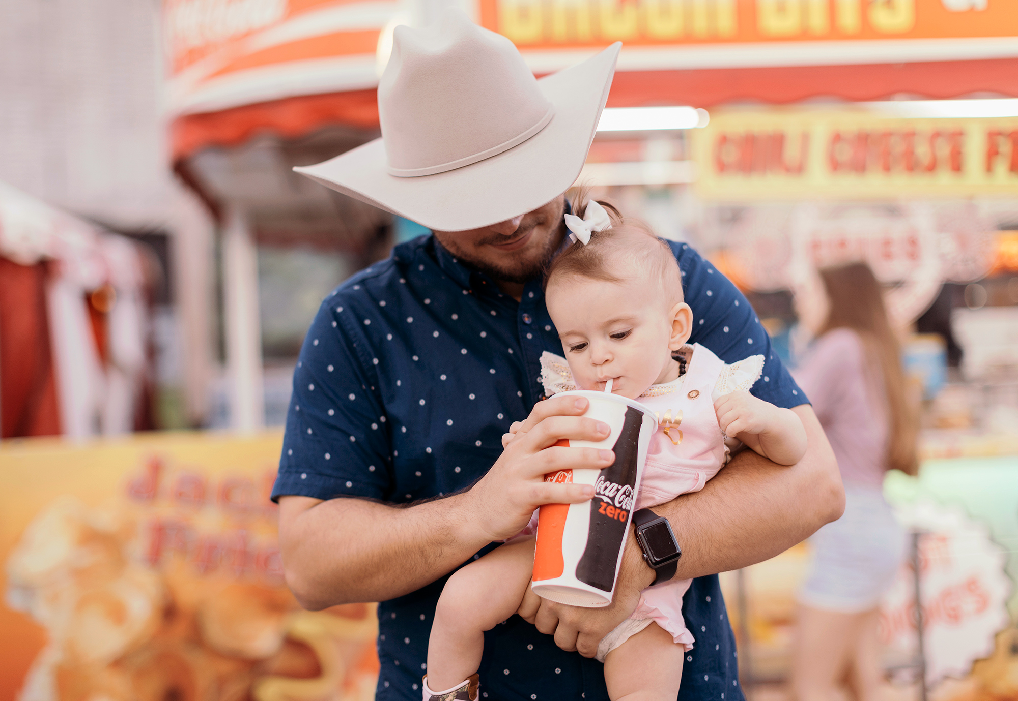 A Houston rodeo carnival attendee lets his baby daughter sip from a soda cup, photographed by Todd Spoth on location at the Houston Rodeo.